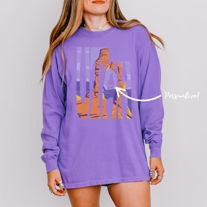 May include: A long-sleeve purple shirt featuring a graphic of a desert landscape with the word "Utah" in vertical stripes. The design includes orange and purple hues, with a white arrow and the word "Personalize!"