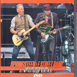Puede incluir: Una caja de DVD que muestra a Bruce Springsteen y Steven Van Zandt actuando en el escenario. Springsteen toca una guitarra eléctrica amarilla, mientras que Van Zandt toca una guitarra eléctrica verde. El texto dice: Springsteen & E Street Band, Land of Hope and Dreams Tour, Olympiastadion Berlin DE, Jun 17 25.