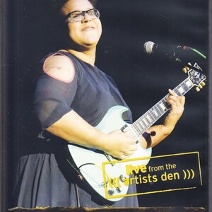 Può includere: Immagine di un'esibizione dal vivo con una persona che suona una chitarra elettrica azzurra. L'immagine include il testo "Alabama Shakes" e "live from the artists den". La persona indossa occhiali e un top blu scuro.