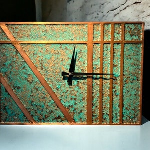 May include: A square clock with a copper-toned frame and a turquoise-colored face. The clock has a black hour and minute hand and a copper-toned design on the face.