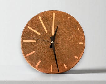 May include: A circular wall clock in a copper tone, featuring black hands and hour indicators. The clock has a speckled texture and a minimalist design. The hour markers are short lines. The clock is on a white surface.
