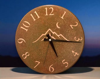 May include: A circular, bronze-toned clock with a mountain range motif. The clock face has white numerals and hands. The clock is set against a blue and purple gradient background.