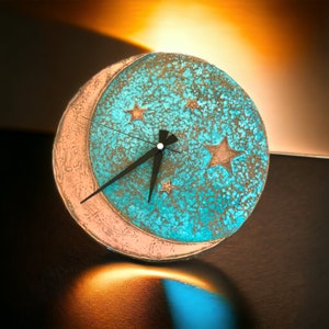 May include: A turquoise and copper clock with a crescent moon design and five stars. The clock hands are black.