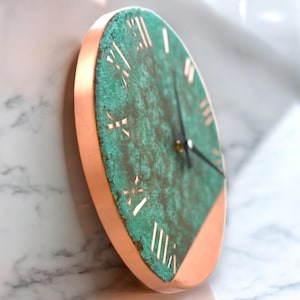 May include: A round wall clock with a copper-colored frame and a verdigris face. The clock face features Roman numerals and black clock hands. The clock is mounted on a white marble surface.