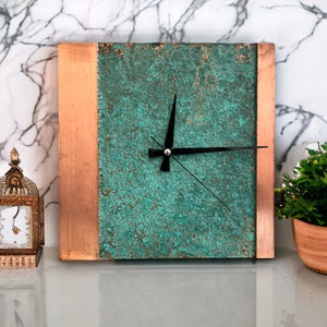 Puede incluir: Reloj cuadrado con borde de cobre y una cara verde azulada. Las manecillas del reloj son negras.