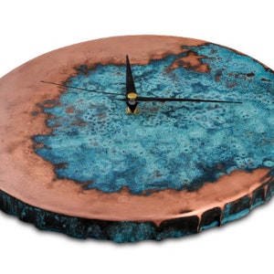 K&ouml;nnte beinhalten: Eine runde Kupferuhr mit einer blauen und gr&uuml;nen Patina. Das Zifferblatt der Uhr hat schwarze Zeiger und eine goldene Mitte.