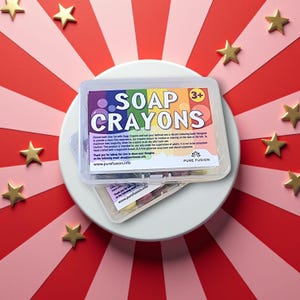 May include: A clear plastic container with a colourful label that reads "SOAP CRAYONS" and "3+". The container holds colourful soap crayons. The container is sitting on a white plate with a red and pink striped background.
