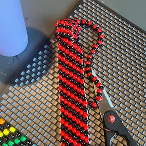 May include: A rectangular beaded cuff bracelet featuring a diagonal striped pattern of alternating red and black plastic pony beads. The bracelet is constructed with a woven design and is positioned next to a pair of black metal scissors.