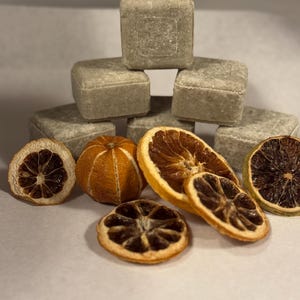 Puede incluir: Una pirámide de seis jabones cuadrados de color verde claro se muestra con rodajas de naranja y limón secas. Las rodajas de cítricos son de color naranja, marrón y amarillo. Los jabones tienen una superficie texturizada.