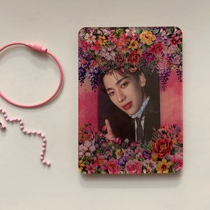 Puede incluir: Una tarjeta de foto rosa y blanca con un borde floral. Un hombre con cabello oscuro aparece en el centro de la tarjeta. Lleva un traje negro y un cuello blanco. Está sonriendo y haciendo un pulgar hacia arriba.