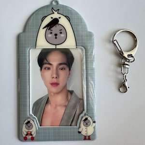 May include: A gray plastic photo card holder with a clear plastic window. The holder has a cartoon image of a bear wearing a white hooded jacket on the top and two similar cartoon bears on the bottom. The holder has a silver metal key chain attached to it.