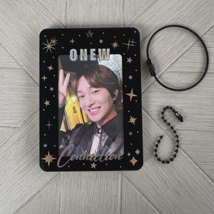 Puede incluir: Portafotos rectangular negro con el nombre "ONEW" y la palabra "Connection" en escritura blanca. El soporte tiene una foto de una persona sonriente. Un llavero de metal negro y una cadena de cuentas están al lado del soporte.
