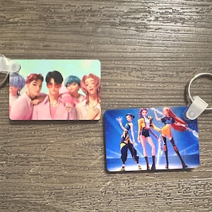 May include: Two rectangular keychains with metal rings. One features a group of people in pink and peach tones. The other shows three figures in black, yellow, and orange against a blue background.