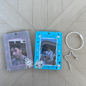 May include: Two photo card holders, one light blue and one gray, with photos inside. The blue holder has a bunny and heart design, while the gray holder has a flower design. A white keychain and a beaded chain are also present.