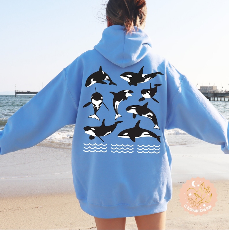 May include: A light blue hoodie with a graphic of multiple black and white orcas swimming in a line. The orcas are surrounded by white waves. The hoodie is being worn by a person standing on a beach.