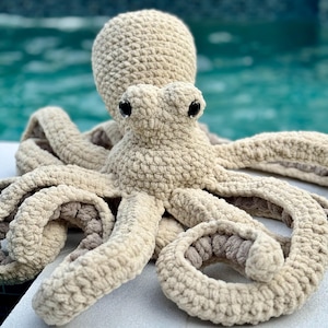 May include: A crocheted octopus plush toy with a beige color and large black eyes. The octopus has eight tentacles and is sitting on a white surface.