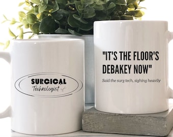 Puede incluir: Dos tazas de cerámica blanca con texto negro. La taza de la izquierda tiene el texto "SURGICAL Technologist" en un óvalo negro con un par de tijeras. La taza de la derecha tiene el texto "IT'S THE FLOOR'S DEBAKEY NOW" y el texto "Said the surg tech, sighing heavily".
