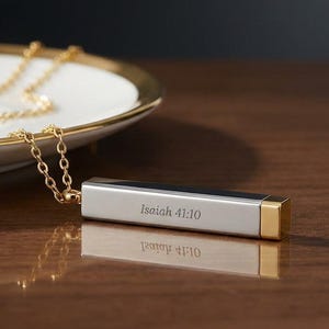 May include: A gold chain necklace with a rectangular pendant. The pendant is silver with gold accents and the text "Isaiah 41:10". The necklace rests on a white plate with a gold rim.