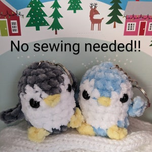 May include: Two plush penguin keyrings, one grey and one light blue, with white bellies and yellow beaks. The penguins are sitting on a white knitted surface. The text "No sewing needed!" is visible in the background.
