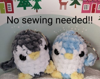 May include: Two plush penguin keychains, one gray and one light blue, with white bellies and yellow beaks. The penguins are sitting on a white knitted surface. The text "No sewing needed!" is visible in the background.