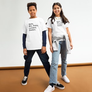Puede incluir: Dos niños con camisetas blancas con los nombres "Belle, Mrs. Potts, Lumière, Gaston" impresos en negro. El niño lleva una camisa azul marino de manga larga debajo de su camiseta y vaqueros azul oscuro. La niña lleva vaqueros azul claro y una sudadera gris.