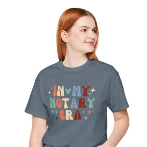 May include: A heather gray t-shirt with the text "IN MY NOTARY ERA" in colorful, retro-style lettering. The design includes hearts, stars, and a vintage font. The shirt is worn by a person in blue jeans.