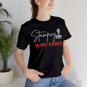 Puede incluir: Camiseta negra con la frase "Stamping is my Cardio" en letras grises y rojas. Un icono de sello gris acompaña el texto. La camiseta es de material suave, ideal para uso casual y aficionados al scrapbooking.