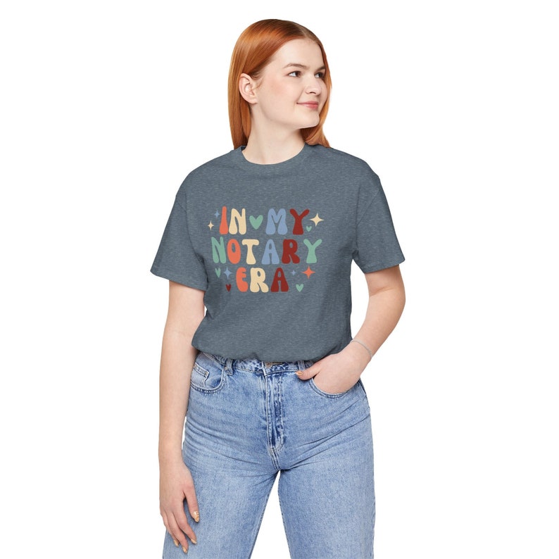 May include: A heather gray t-shirt with the text "IN MY NOTARY ERA" in colorful, retro-style lettering. The design includes hearts, stars, and a vintage font. The shirt is worn by a person in blue jeans.