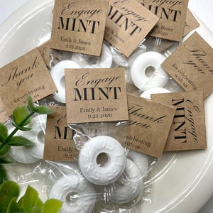 May include: White mint candies in clear plastic bags, arranged on a white plate. Each bag has a small, square, brown tag that reads "Engage MINT" with a date and names. Some tags say "Thank you".