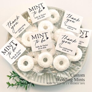 May include: A collection of white ring-shaped mint candies individually wrapped in clear plastic, each topped with a square paper tag. The tags feature black text reading MINT to be, Sally & Taylor 10.24.2026, or Thank you for celebrating with us.