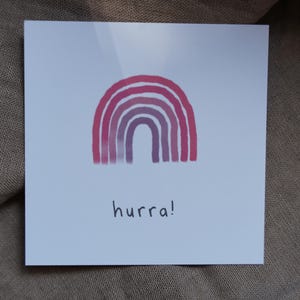 May include: A square white card with a watercolor rainbow in shades of pink and purple. The word "hurra!" is written in black script below the rainbow. The card is on a neutral-colored fabric background.