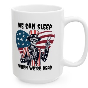 May include: White ceramic coffee mug with a graphic of a skeleton dressed as Uncle Sam. The skeleton is holding a peace sign and is standing in front of a heart-shaped American flag. The text "We can sleep when we're dead" is printed on the mug.
