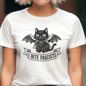 Puede incluir: Camiseta blanca con una ilustración de un gato negro con alas de murciélago y una capa. El gato está sentado sobre una pancarta con el texto "I BITE FASCISTS". El diseño es un gráfico en blanco y negro.
