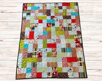May include: A patchwork quilt with a black border, featuring a variety of colorful squares. The squares include patterns of red, green, blue, and brown, with holiday-themed designs like houses and stripes. The quilt is displayed on a light wood surface.