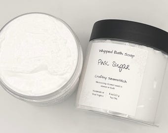 May include: Two clear plastic jars filled with white whipped bath soap. One jar is open, showing the textured cream, while the other is closed with a black lid and a label reading Whipped Bath Soap, Pink Sugar, Craftsy Seamstitch, 8 oz/227 g.