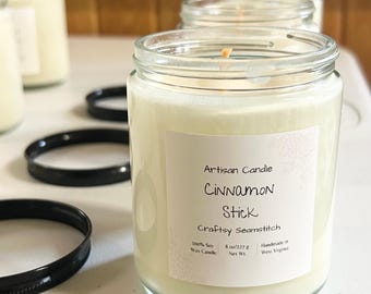 May include: Several glass jar candles with black lids are visible. The front candle has a label that reads "Artisan Candle Cinnamon Stick Craftsy Seamstitch 100% Soy Wax Candle 227 g Net Wt. Handmade in West Virginia."