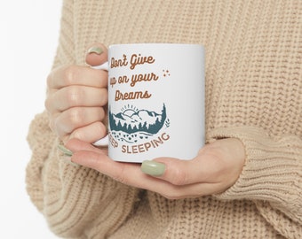 Könnte beinhalten: Eine weiße Keramiktasse mit einem braunen und blauen Design. Die Tasse zeigt eine Bergszene mit Sonnenaufgang und dem Text "Don't Give Up On Your Dreams Keep Sleeping".