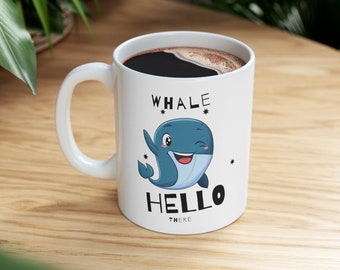 Könnte beinhalten: Weiße Keramiktasse mit schwarzem Henkel. Die Tasse zeigt eine blaue Wal-Karikatur mit einem Augenzwinkern und den Text "Whale Hello There" in Schwarz.