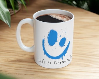 Könnte beinhalten: Weiße Keramik-Kaffeetasse mit einem blauen Smiley und dem Text "Life is Brew-tiful".
