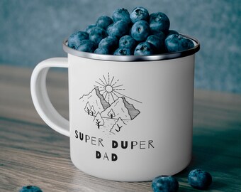 Könnte beinhalten: Weiße Emaille-Campingtasse mit einer Schwarzweißillustration von Bergen, einer Sonne und Bäumen. Der Text "Super Duper Dad" ist unter der Illustration gedruckt. Die Tasse ist mit Blaubeeren gefüllt.