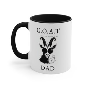 May include: White ceramic coffee mug with a black handle and black rim. The mug has a black and white illustration of a goat wearing sunglasses and the text "G.O.A.T. Greatest of All Time Dad".