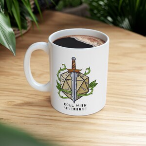 May include: White ceramic mug with a black handle. The mug has a graphic of a sword through a 20-sided die with vines. The text "Roll with Adventure" is printed below the graphic.