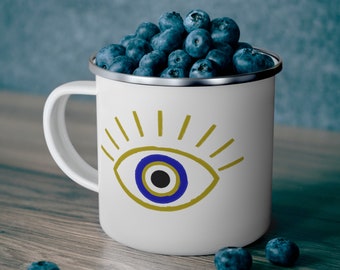 Könnte beinhalten: Eine weiße Emailletasse mit einem blauen und goldenen bösen Auge. Die Tasse ist mit Blaubeeren gefüllt.