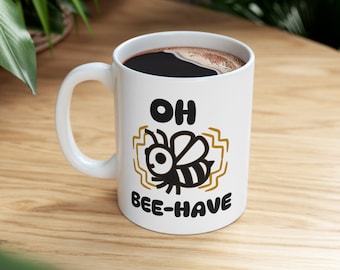 Könnte beinhalten: Weiße Keramik-Kaffeetasse mit einem schwarzen und goldenen Design. Die Tasse zeigt eine schwarze Bienenillustration und den Text "OH BEE-HAVE".
