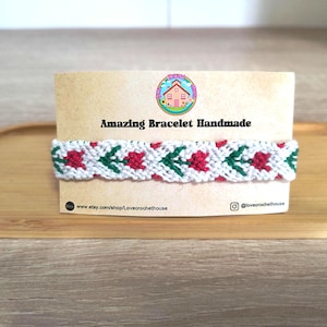 Puede incluir: Una pulsera blanca hecha a mano con un patrón repetido de flores rojas y hojas verdes. La pulsera se muestra en una tarjeta marrón con el texto "Amazing Bracelet Handmade". La URL de la tienda Etsy es visible.