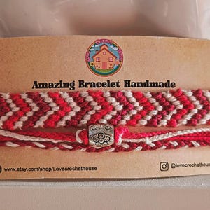 May include: Two handmade bracelets on a tan card labeled Amazing Bracelet Handmade. The top bracelet features a red and white chevron pattern. The bottom bracelet is a thin red and white twisted cord with a silver bead engraved with SIS.