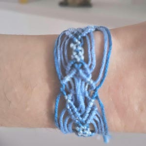 May include: A handmade macrame bracelet in shades of blue, featuring a woven central design and dangling strands. The bracelet is worn on a wrist, showcasing the intricate knotting and color variations.