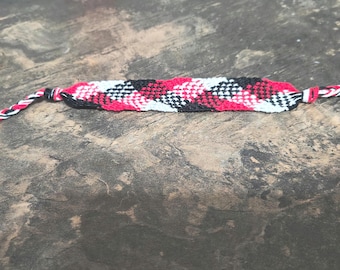May include: A handmade friendship bracelet featuring a woven pattern of red, black, and white threads. The bracelet is displayed on a textured, brown surface. The ends of the bracelet are braided with red, black, and white threads.