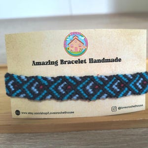 Puede incluir: Una pulsera hecha a mano con un patrón geométrico en negro, azul claro y rojo. La pulsera se muestra en una tarjeta beige con el texto "Amazing Bracelet Handmade" y un logotipo colorido. La URL de la tienda Etsy es visible.