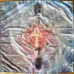 May include: A square cloth with a celestial design featuring a pink and white rose-like pattern, set against a blue and purple galaxy backdrop. An Ankh symbol, crystals, and small, round stones are arranged on the cloth. Likely used for spiritual purposes.